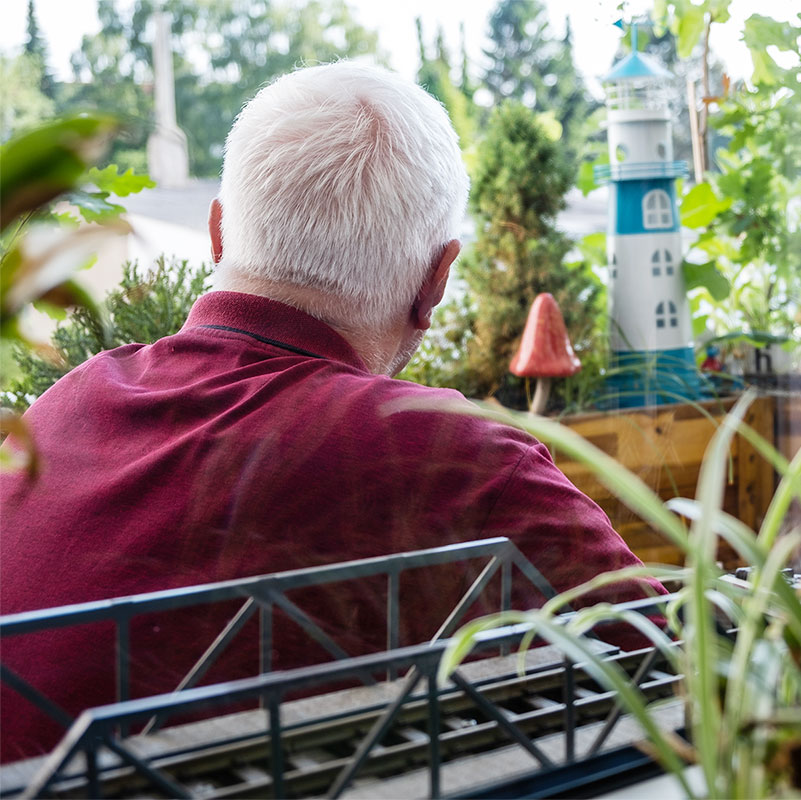 Rückansicht eines Mannes mit weißen Haaren, der auf seinem Balkon sitzt und in die Ferne schaut